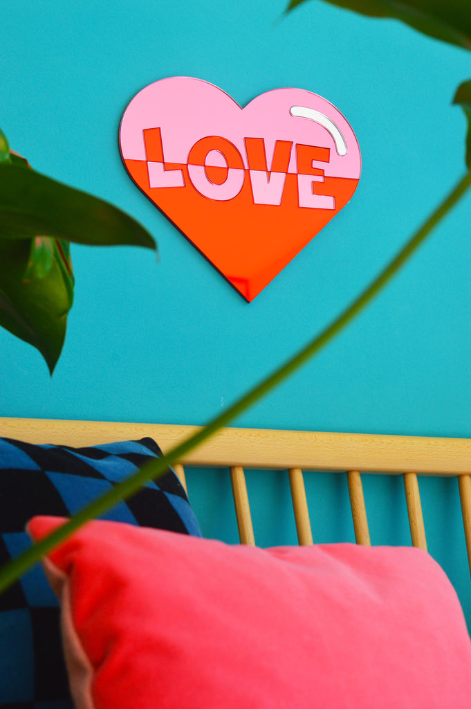 This image features the Love Heart Wall Art, a beautiful piece displayed on a vibrant turquoise wall above a seat. The wall art, crafted from acrylic mirrors in bright pink and red hues, showcases the word 'love.' The contrasting colors and elegant design make it a focal point, adding warmth and charm to the space. This description is tailored for those using screen readers or with limited bandwidth, providing a vivid mental image of the Love Heart Wall Art in its setting