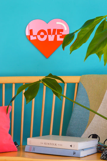 This image features the Love Heart Wall Art, a beautiful piece displayed on a vibrant turquoise wall above a seat. The wall art, crafted from acrylic mirrors in bright pink and red hues, showcases the word 'love.' The contrasting colors and elegant design make it a focal point, adding warmth and charm to the space. This description is tailored for those using screen readers or with limited bandwidth, providing a vivid mental image of the Love Heart Wall Art in its setting